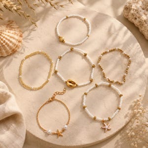 May include: A set of six bracelets featuring white seed beads, gold-toned metallic beads, and charms including a central cowrie shell, a starfish, and a small shell. The collection includes elastic beaded strands and a gold-toned metal chain bracelet.