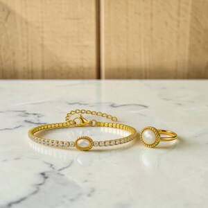 May include: A gold-toned bracelet and a matching ring, both featuring a central oval white stone surrounded by a beaded border. The bracelet has a chain of small clear stones and a lobster clasp, while the ring has a simple gold band.