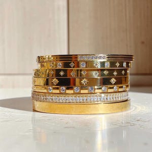 Może przedstawiać: Stos sześciu złotych metalowych bransoletek typu bangle. Bransoletki mają różne wzory, w tym przezroczyste okrągłe kamienie, grawerunki w kształcie gwiazd oraz opaskę w całości wysadzaną małymi, prostokątnymi, przezroczystymi kamieniami.