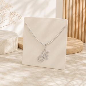Puede incluir: Un collar tipo tenis de tono plateado con un colgante en forma de letra A cursiva. Tanto la cadena como el colgante están incrustados con pequeñas piedras claras y brillantes que captan la luz, creando un efecto resplandeciente en la joya.
