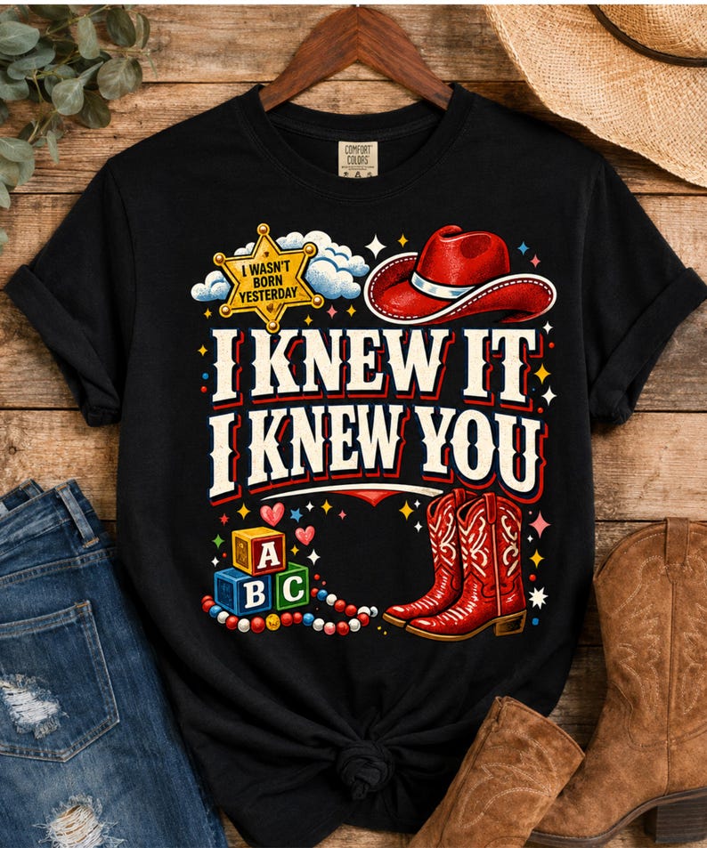 Puede incluir: Una camiseta negra de manga corta con un estampado gr&aacute;fico que incluye una estrella de sheriff amarilla con la inscripci&oacute;n I WASN'T BORN YESTERDAY, un sombrero de vaquero rojo, botas de vaquero rojas y bloques de letras. El texto dice I KNEW IT I KNEW YOU.