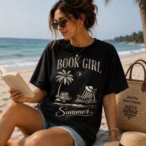 Op de afbeelding: Een zwart t-shirt met korte mouwen en een witte grafische print met de tekst BOOK GIRL en Summer, een palmboom, een stapel boeken en een strandstoel. Het shirt wordt gecombineerd met een lichtblauwe spijkerbroek en een rieten tas met tekst.