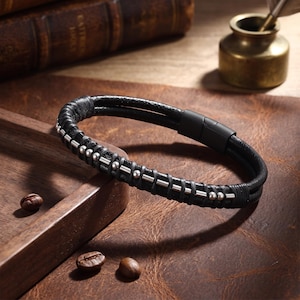 May include: A black leather bracelet featuring a double-strand design with silver-toned metal beads and cylindrical spacers arranged in a pattern along the cord. The bracelet is secured with a matte black cylindrical magnetic clasp closure.