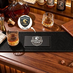 Può includere: Un tappetino da bar rettangolare in gomma nera con superficie testurizzata a rilievo e una stampa grafica bianca centrale raffigurante una testa d'aquila all'interno di uno scudo con il testo Golden Eagle Lounge su legno.