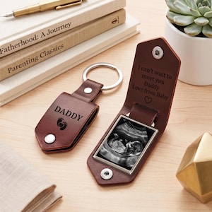May include: Two brown leather keychains with silver metal rings. One is closed, showing the word DADDY and baby footprints. The other is open, revealing a square frame containing a black and white ultrasound image and the text I can't wait to meet you Daddy Love from Baby with a heart symbol.