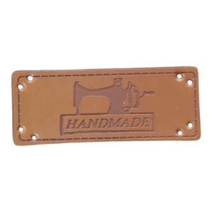 May include: A rectangular tan leather label featuring an embossed graphic of a vintage sewing machine above the word HANDMADE. The label has a stitched border design and includes four small pre-punched holes at the corners for attachment.