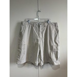May include: Beige cargo shorts hanging on a clear plastic hanger. The shorts feature two front pockets, two side cargo pockets, and two back pockets. The shorts are made of a sturdy material.