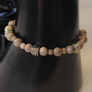 Può includere: Un braccialetto di perline rotonde color beige con una superficie strutturata. Il braccialetto include dettagli metallici color argento. Il braccialetto è esposto su un supporto nero.