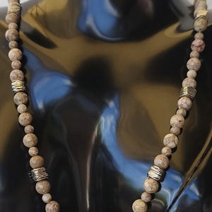Può includere: Collana di perline con perline rotonde marrone chiaro dall'aspetto strutturato. Distanziatori in metallo color argento sono intercalati. La collana è esposta su un manichino nero. Gioielleria.