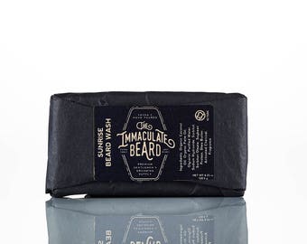 May include: A black bar of soap wrapped in black paper with a label that reads "The Immaculate Beard" and "Sunrise Beard Wash". The label also lists ingredients including organic coconut oil, organic palm oil, glycerin, purified water, sodium hydroxide, sorbitol, sorbitan oleate, soybean protein, shea butter, activated charcoal, and fragrance. The soap is 4.25 oz (120 g).
