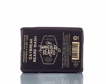 May include: A black bar of soap with gold lettering that reads "The Immaculate Beard Daybreak Beard Wash" with a list of ingredients and a net weight of 4 oz. The soap is labeled with a spearmint, eucalyptus, lavender, and clary sage scent.