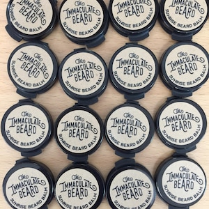 Puede incluir: Una colección de recipientes negros de bálsamo para barba. Cada recipiente tiene una etiqueta circular con el texto "The Immaculate Beard Sunrise Beard Balm" en una fuente estilizada. Los recipientes están dispuestos en forma de cuadrícula.