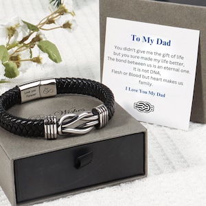 Op de afbeelding: Een zwarte gevlochten leren armband met een zilverkleurig metalen knoop als middelpunt en een magnetische sluiting gegraveerd met "Love you Forever" en een oneindigheidssymbool, gepresenteerd met een "To My Dad" kaart en grijze geschenkdoos.