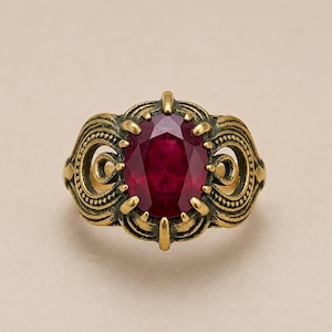 Könnte beinhalten: Ein Ring im antiken Stil mit einem großen, oval geschliffenen Rubin-Edelstein. Der Ring hat eine detaillierte, verzierte Fassung in Goldtönen mit wirbelnden Mustern und filigranen Akzenten. Der Rubin hat eine tiefrote Farbe, vor neutralem Hintergrund.