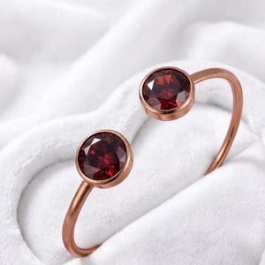 Puede incluir: Una pulsera de brazalete abierto en oro rosa con dos piedras granate de color rojo intenso, redondas y facetadas, engastadas en biseles circulares en cada extremo. La banda de metal tiene un acabado liso y pulido con un diseño abierto.
