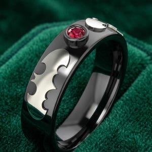 May include: A black band ring featuring a silver-toned bat-shaped inlay and a single round, faceted red gemstone set in a raised black bezel. The ring has a polished finish and a smooth, rounded profile, showcasing a distinct bat silhouette design.