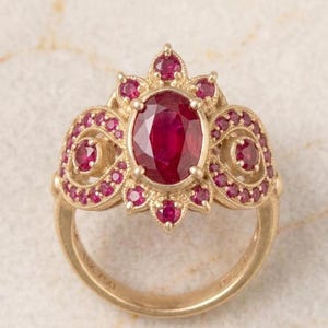 May include: A gold ring featuring a large, oval-cut ruby center stone, surrounded by smaller rubies in a floral and swirl design. The ring is marked "18K 750". The ring's design is ornate and detailed, with a vintage or antique aesthetic.