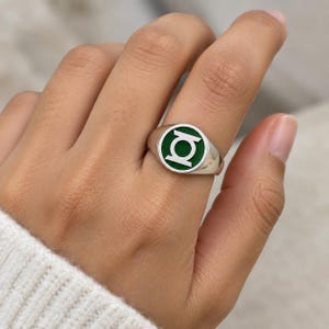 May include: A silver-toned signet ring featuring a circular face with a green enamel inlay. The center of the ring displays a silver-colored Green Lantern symbol, which consists of a circle with a vertical line and two horizontal bars.