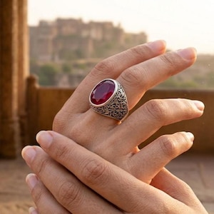 Puede incluir: Un anillo de plata con una piedra preciosa ovalada de color rojo intenso. El anillo tiene un diseño ornamentado y grabado alrededor de la piedra. El anillo está en una mano, con un fondo borroso de un edificio y luz natural.