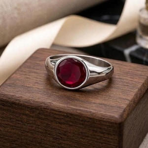 Op de afbeelding: Een zilveren ring met een grote, ronde, dieprode edelsteen. De ring ligt op een donkerbruin houten blok, met een lint en een kleine fles op de achtergrond. Het ontwerp van de ring is eenvoudig en elegant.