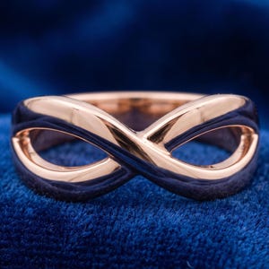May include: A polished rose gold ring featuring a continuous infinity symbol design. The band has a smooth, reflective surface that curves into two symmetrical loops, creating a fluid and interconnected shape that forms the center of the ring.