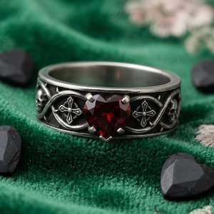 May include: A silver-toned metal ring featuring a central heart-shaped dark red gemstone. The band is decorated with an embossed pattern of skulls, crosses, and thorny vine motifs that wrap around the entire circumference of the jewellery piece.