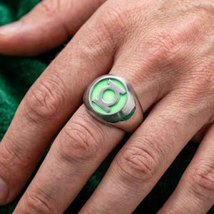 May include: A silver-toned metal ring featuring a circular face with a glowing light green inlay. The center of the inlay displays the iconic Green Lantern symbol in silver metal, creating a distinct contrast against the luminous green background.
