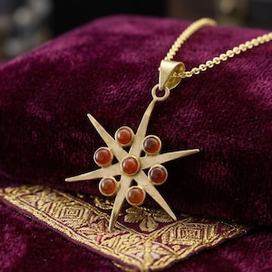 May include: A gold-colored starburst pendant necklace with a delicate chain. The starburst features eight pointed rays and is adorned with seven round, reddish-orange gemstones. The necklace rests on a deep purple velvet surface.