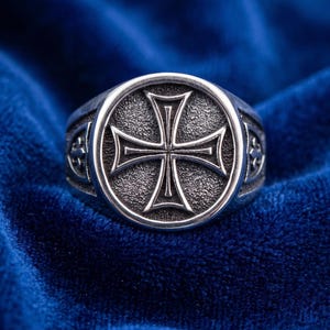 Könnte beinhalten: Ein silberfarbener Ring mit einer kreisförmigen Vorderseite, die ein erhabenes Malteserkreuz-Design vor einem texturierten, dunklen Hintergrund zeigt. Die Ringschiene weist an den Seiten zusätzliche dekorative Details auf und hat ein poliertes Finish.