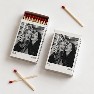 May include: Two rectangular matchboxes with black and white photographic prints of two smiling people holding soda cans. The boxes feature the text E.S and 2006. One box is open, revealing wooden matches with red tips. Three loose matches are nearby.