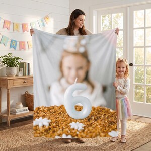 Op de afbeelding: Een rechthoekige fleece deken met een bedrukte foto van een verjaardagstaart met daarop een grote, lichtblauwe kaars in de vorm van het cijfer zes. De afbeelding toont een wazig persoon op de achtergrond en een textuurrijke, goudbruine taartoppervlak.