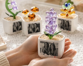 Könnte beinhalten: Ein quadratischer weißer Keramikübertopf mit einer violetten Glasblumenskulptur und hellem Kies. Auf der Vorderseite des Übertopfs befindet sich ein schwarz-weißes Foto von zwei Personen und der Text "Whatever Forever" mit einem kleinen roten Herz.