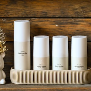 Op de afbeelding: Een collectie Fresh lichaamsverzorgingsproducten, waaronder body milk, handbalsem, voetbalsem en deodorant, in witte en beige cilindrische flessen. De producten worden gepresenteerd op een beige dienblad.