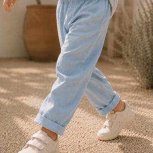 Può includere: Pantaloni azzurri con elastico in vita e orli risvoltati, abbinati a sneakers bianche basse con doppia chiusura a strappo. I pantaloni hanno una vestibilità rilassata e sono indossati con una camicia a righe azzurre e bianche.