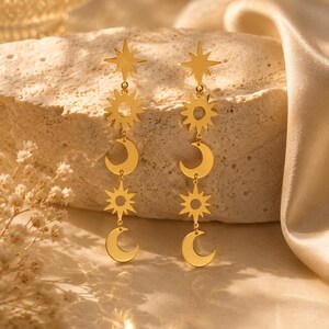 Puede incluir: Pendientes celestiales dorados con diseños de estrella, sol y luna creciente. Los pendientes son largos y colgantes, con cada elemento conectado por pequeños bucles. Se exhiben sobre una superficie texturizada de color claro.