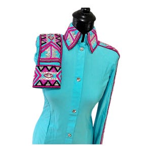 May include: A bright turquoise long-sleeve shirt featuring a structured collar and cuffs adorned with intricate geometric patterns in hot pink, black, and silver. The design includes rows of sparkling crystals and clear rhinestones throughout.