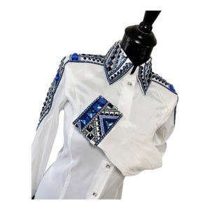 May include: A white long-sleeve button-up shirt featuring intricate embellishments on the collar, shoulders, and cuffs. The design includes geometric patterns of blue, silver, and black crystals and studs, creating a detailed decorative border.