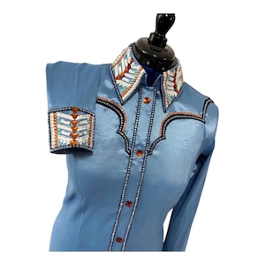 May include: A light blue long-sleeve shirt featuring a pointed collar and cuffs decorated with geometric patterns of white, light blue, and orange crystals. The front placket and yoke are accented with rows of small clear crystals and orange buttons.