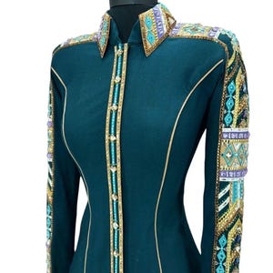 May include: Teal-colored button-down shirt with intricate embellishments. The shirt features gold trim, turquoise and gold beading, and decorative patterns on the collar and sleeves. The long-sleeved shirt is displayed on a mannequin.