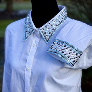 May include: White button-down shirt with a decorative collar and cuffs. The collar and cuffs are embellished with a pattern of clear and blue rhinestones and silver studs. The shirt is displayed on a black mannequin.
