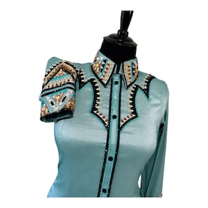 May include: A light blue long-sleeve shirt featuring a pointed collar and front button placket. The garment is decorated with intricate geometric patterns of clear, gold, and dark crystals, accented by black trim along the collar and chest.