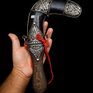 Op de afbeelding: Een gebogen dolk met een donkerhouten handvat en een zwarte schede met ingewikkeld zilveren filigraanwerk. De schede is versierd met een felrood gevlochten koord dat is bevestigd aan twee kleine metalen ringen aan de zijkanten van de zilveren houder.