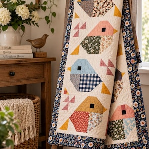 May include: A rectangular quilt featuring a repeating pattern of stylized birds in various colors and floral or gingham fabric patches. The quilt has a cream-colored background and is bordered by a dark blue fabric with a white and yellow daisy print.