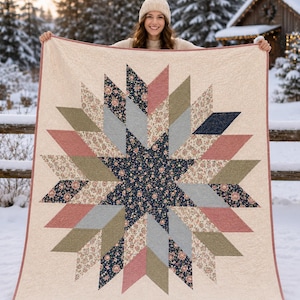 Pode incluir: Um edredom bege com um grande design de estrela. A estrela é composta por várias formas geométricas em tons de rosa, azul, verde e cinza, com estampas florais. O edredom tem uma borda rosa e é exibido ao ar livre em um ambiente de neve.