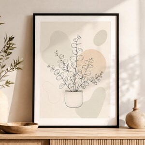 Peut inclure: Une affiche encadrée représentant un dessin au trait noir d'une plante d'eucalyptus en pot. L'arrière-plan se compose de formes abstraites douces dans des tons beige, vert pâle et pêche, le tout dans un cadre noir simple sur une surface en bois.