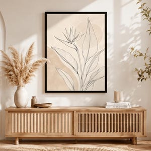 Peut inclure: Une impression d'art mural encadrée présentant un dessin au trait noir d'une fleur oiseau du paradis avec de grandes feuilles sur un fond aquarelle beige et crème. L'œuvre est entourée d'un cadre noir fin et affiche des éléments botaniques.
