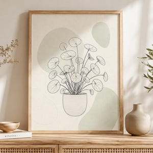 Peut inclure: Une impression d'art encadrée représentant un dessin au trait noir d'une plante Pilea dans un pot texturé, sur un fond crème avec des formes organiques vert sauge. L'œuvre est présentée dans un cadre en bois clair sur une surface en bois.