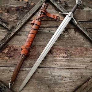 Può includere: Una spada in acciaio con guardia a croce e pomolo circolare, posizionata accanto a un fodero in pelle marrone. Il fodero presenta una lunga cinghia di cuoio avvolta lungo la sua lunghezza, fissata con una fibbia metallica e una punta in metallo.