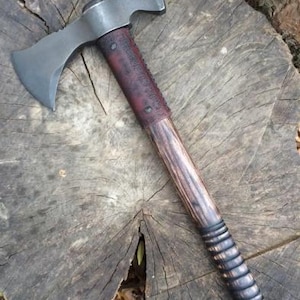 Op de afbeelding: Een dubbelzijdige tomahawk met een donkergrijze metalen kop met twee scherpe bladen. De steel is gemaakt van hout met een donkerbruine lederen wikkel die bij de kop is vastgezet met metalen klinknagels en heeft een reeks uitgesneden ribbels.