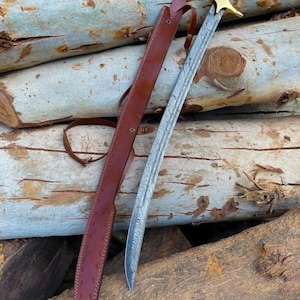 Puede incluir: Una espada con hoja de acero Damasco estampada, una guarda dorada y un mango negro con remaches plateados. La espada viene acompañada de una funda de cuero marrón que cuenta con una correa de cuero a juego y cierres de presión metálicos.